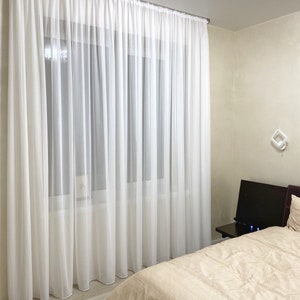 May include: White sheer curtains hang in a bedroom with a beige bedspread and a dark brown nightstand. The curtains are hung on a silver curtain rod.