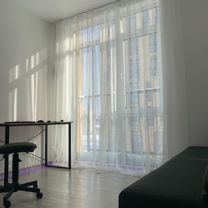 May include: A white room with a large window covered in sheer white curtains. A black desk with a black chair sits in the corner of the room. A gray couch is in the foreground.