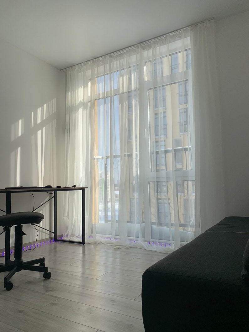 May include: A white room with a large window covered in sheer white curtains. A black desk with a black chair sits in the corner of the room. A gray couch is in the foreground.