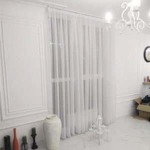 May include: White sheer curtains hang in a room with white walls and a white tiled floor. A white ceramic vase with a decorative design sits on the floor in front of the curtains. A white metal candelabra with six candle holders sits on the floor in front of the curtains.