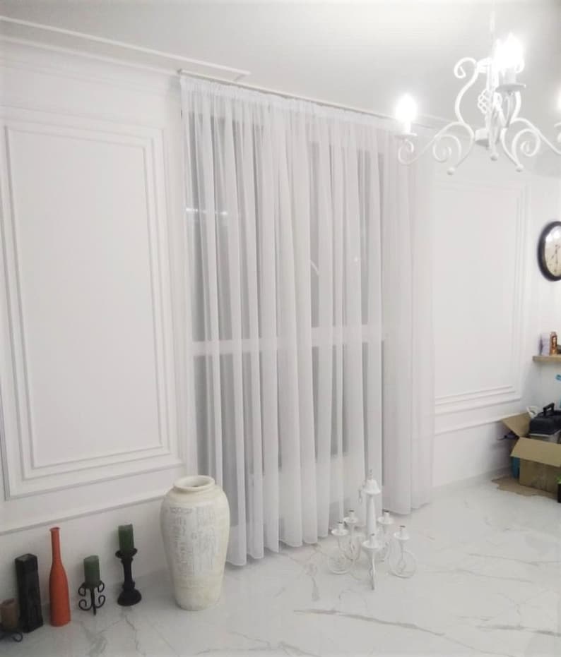 May include: White sheer curtains hang in a room with white walls and a white tiled floor. A white ceramic vase with a decorative design sits on the floor in front of the curtains. A white metal candelabra with six candle holders sits on the floor in front of the curtains.