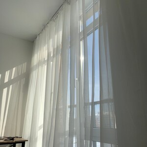 May include: White sheer curtains hang in a window, allowing sunlight to stream through the fabric. The curtains are gathered at the top and hang in soft folds.