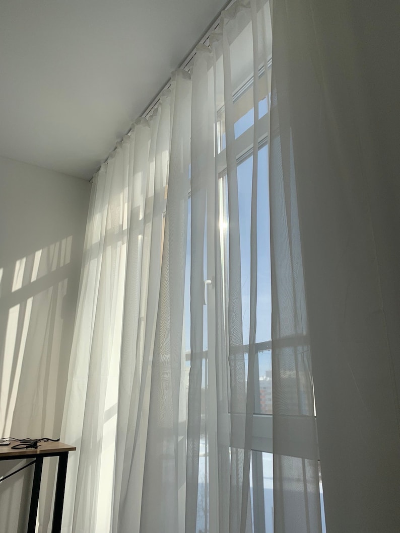 May include: White sheer curtains hang in a window, allowing sunlight to stream through the fabric. The curtains are gathered at the top and hang in soft folds.