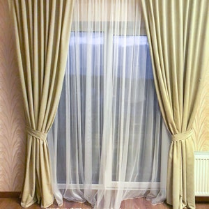 May include: A pair of beige curtains with a white sheer curtain behind them. The curtains are tied back with a knot and the sheer curtain is flowing.