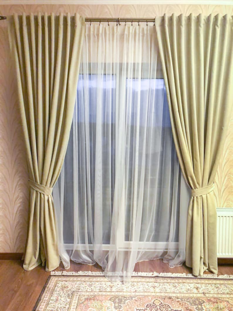 May include: A pair of beige curtains with a white sheer curtain behind them. The curtains are tied back with a knot and the sheer curtain is flowing.