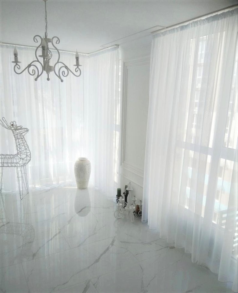 May include: White sheer curtains hang in a room with white tile floors and a white ceramic vase. A white chandelier hangs from the ceiling.