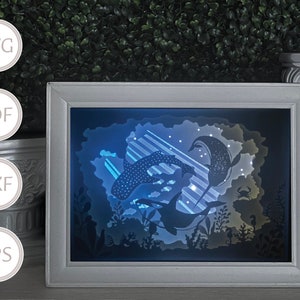 May include: A white frame with a blue and white paper cut-out design of a whale and a manta ray swimming in the ocean. The design is lit from behind, creating a glowing effect.