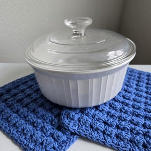 May include: A white ceramic baking dish with a glass lid, sitting on two blue crocheted pot holders.