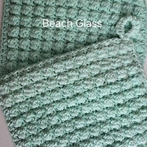 May include: Two crocheted dishcloths in a light green color. The dishcloths have a textured, bumpy surface. The top dishcloth has a loop handle. The text "Beach Glass" is written on the top dishcloth.