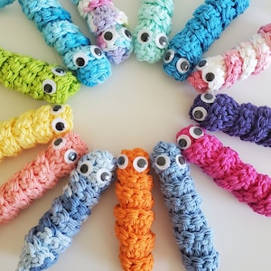 May include: A collection of colorful crocheted worm toys with googly eyes. The worms are arranged in a circle on a white surface.
