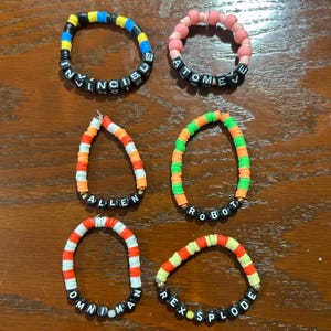 May include: Six circular bracelets made of colorful flat clay beads and black letter beads. The bracelets feature the text INVINCIBLE, ATOMEJE, ALLEN, ROBOT, OMNI-MAN, and REX-SPLODE. The beads are arranged in various color patterns including blue, yellow, black, pink, orange, green, white, and red.