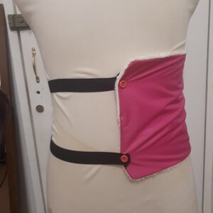 May include: A pink fabric belt with two black elastic straps and two orange buttons. The belt is worn on a white mannequin.