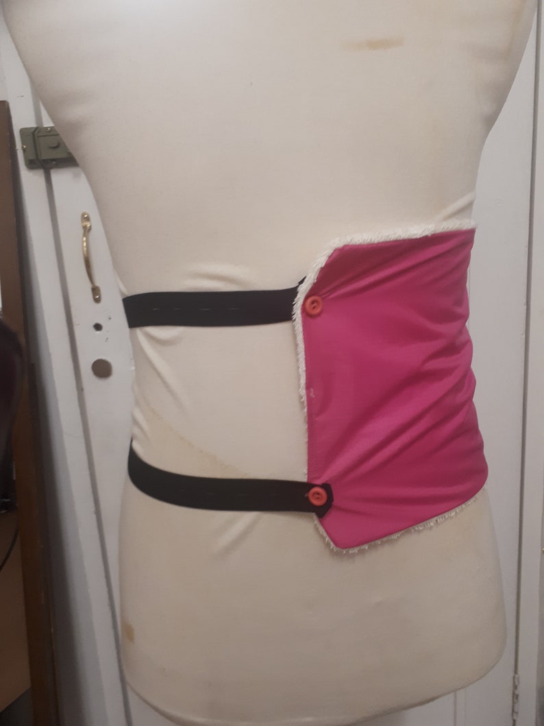 May include: A pink fabric belt with two black elastic straps and two orange buttons. The belt is worn on a white mannequin.