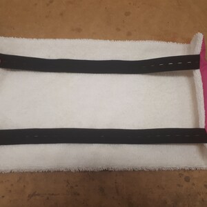 May include: White towel with two black straps and pink edging. The straps have pink buttons on each end.