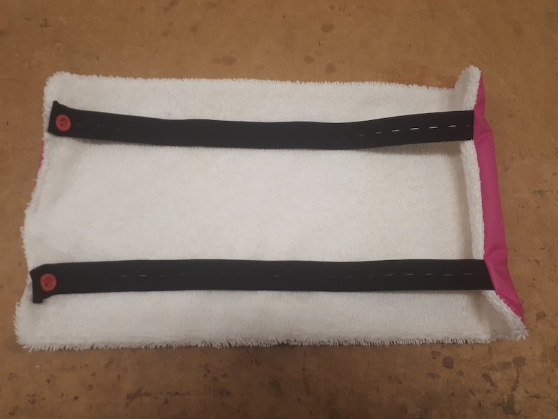May include: White towel with two black straps and pink edging. The straps have pink buttons on each end.