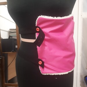 May include: A pink fabric pouch with a white trim and two orange buttons. The pouch is attached to a black elastic band.