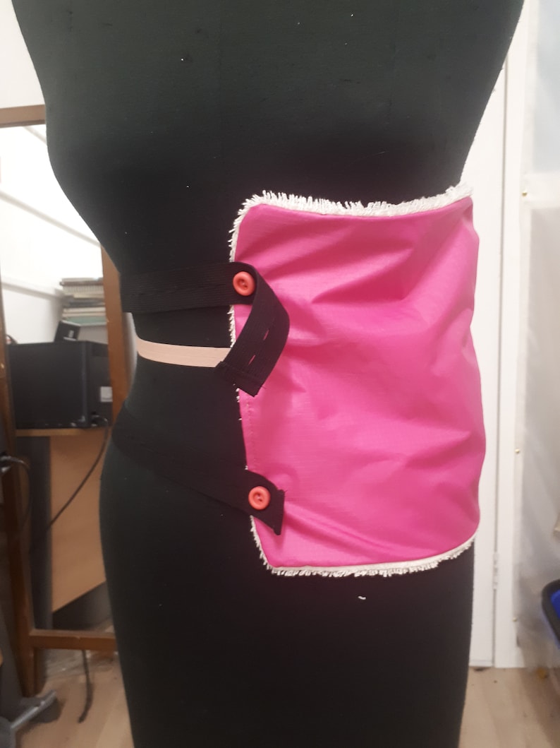 May include: A pink fabric pouch with a white trim and two orange buttons. The pouch is attached to a black elastic band.