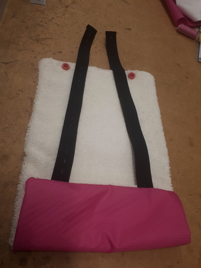 May include: A white, fluffy, rectangular pouch with two black straps and pink buttons. The pouch has a pink fabric lining.