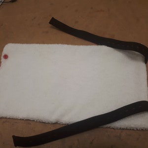 May include: White, fluffy, rectangular pad with two red buttons on the left side. Two black straps are attached to the pad, one on each side.