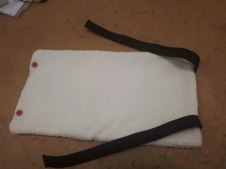 May include: White, fluffy, rectangular pad with two red buttons on the left side. Two black straps are attached to the pad, one on each side.