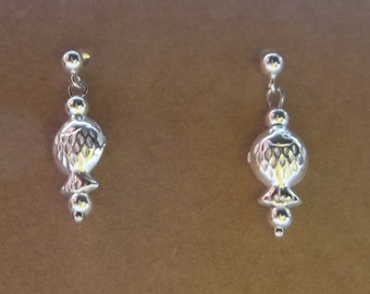 May include: A pair of silver-colored earrings. Each earring features a small ball stud, a chain, a round, textured fish-shaped pendant, and a small ball at the bottom. The earrings are displayed against a brown background.
