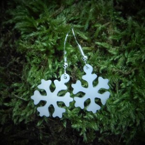 Könnte beinhalten: Ein Paar weiße Schneeflocken-Ohrringe mit silberfarbenen Haken. Die Ohrringe sind vor einem Hintergrund aus grünem Moos platziert.