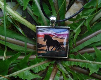 May include: Square pendant with a black horse silhouette against a colorful sunset backdrop. The pendant is set in a silver-colored frame and hangs from a black cord. The background features green foliage.