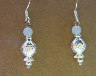 May include: A pair of silver-toned fish earrings. Each earring features a fish-shaped charm with etched details, a pale blue bead, and a curved ear wire. The earrings are displayed against a brown background.