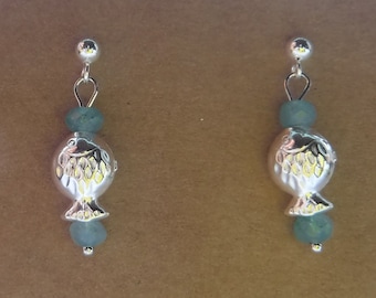 May include: A pair of silver-toned fish earrings with light blue beads. The fish-shaped charms have etched details and dangle from a small silver ball at the top. The earrings are displayed against a brown background.