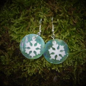 Könnte beinhalten: Ein Paar schillernde runde Ohrringe mit einem weißen Schneeflocken-Design. Die Ohrringe haben silberfarbene Haken und sind vor einem moosgrünen Hintergrund platziert. Ein winterliches Accessoire.
