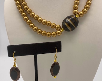 May include: A gold-colored beaded necklace with a large, oval, brown and cream-colored stone pendant. Matching earrings with brown and black oval stones and gold-colored accents are also displayed. The jewelry set is presented on a white mannequin.