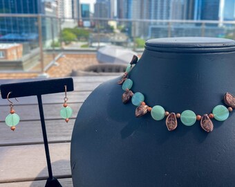May include: A necklace and earring set displayed on a black bust. The necklace features round, light green beads interspersed with copper-colored leaf-shaped charms and small brown beads. The earrings have matching green beads and copper accents.