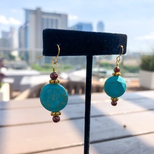 May include: A pair of turquoise and gold earrings. Each earring features a large, round, turquoise bead, accented with gold-colored beads and small, dark red beads. The earrings hang from gold-colored ear wires.