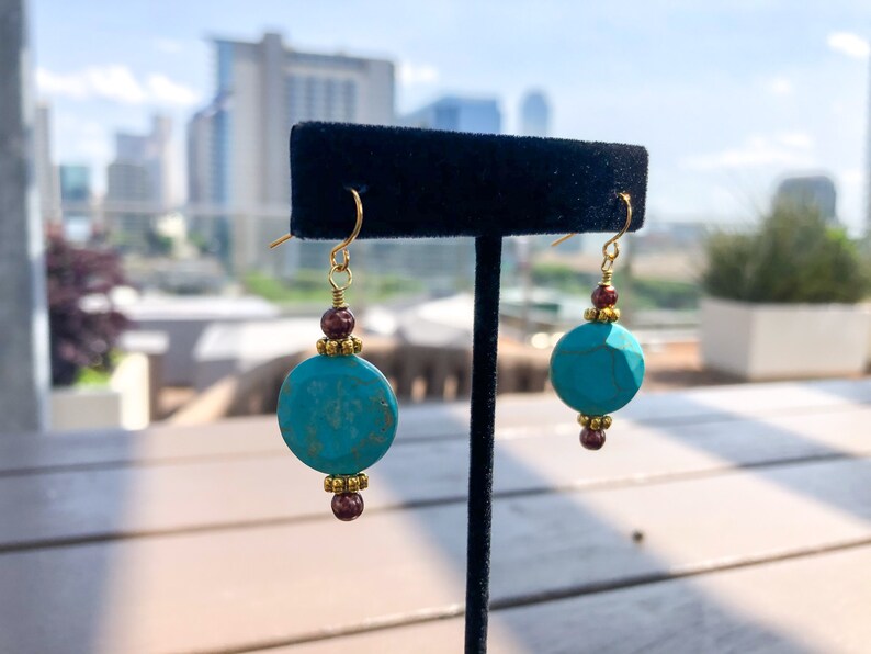 May include: A pair of turquoise and gold earrings. Each earring features a large, round, turquoise bead, accented with gold-colored beads and small, dark red beads. The earrings hang from gold-colored ear wires.
