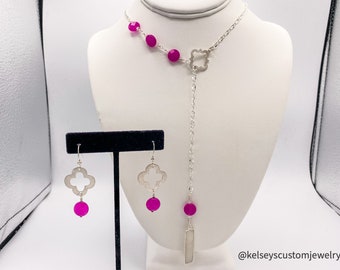 May include: A silver-toned necklace and matching earrings set. The necklace features a clover-shaped pendant and a chain with fuchsia-colored beads. The earrings have a clover design with a fuchsia bead. The jewelry is displayed on a white mannequin.