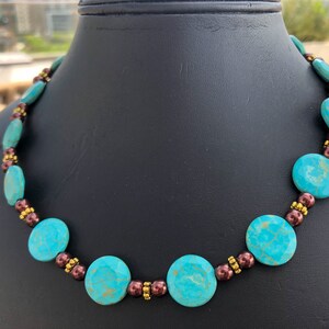 May include: A turquoise beaded necklace with brown and gold accents. The necklace features large, round turquoise beads interspersed with smaller brown and gold beads. The necklace is displayed on a black mannequin.