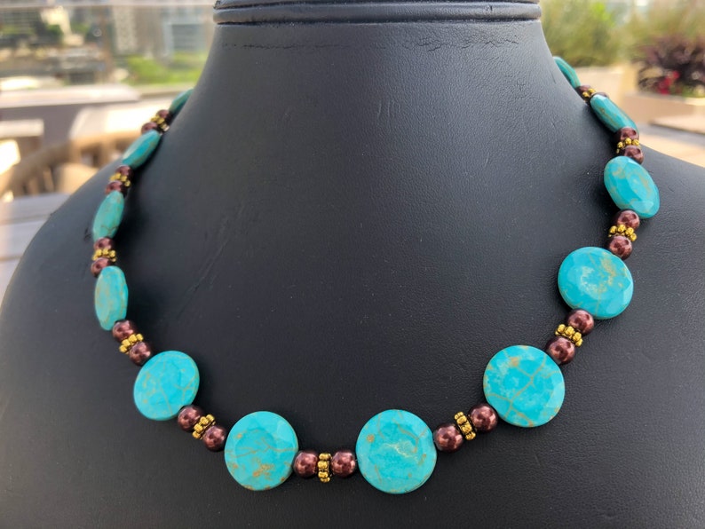 May include: A turquoise beaded necklace with brown and gold accents. The necklace features large, round turquoise beads interspersed with smaller brown and gold beads. The necklace is displayed on a black mannequin.