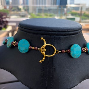 May include: A turquoise and gold necklace displayed on a black mannequin. The necklace features large, faceted turquoise beads interspersed with smaller, dark red beads. A gold-colored clasp secures the necklace.