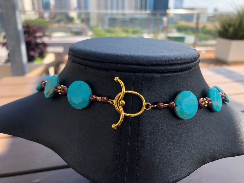 May include: A turquoise and gold necklace displayed on a black mannequin. The necklace features large, faceted turquoise beads interspersed with smaller, dark red beads. A gold-colored clasp secures the necklace.