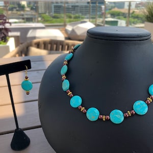 May include: A turquoise necklace and matching earrings set. The necklace features large, round turquoise beads interspersed with smaller brown and gold beads. The earrings have a similar design, with turquoise beads and gold hooks. The jewelry is displayed on a black mannequin.