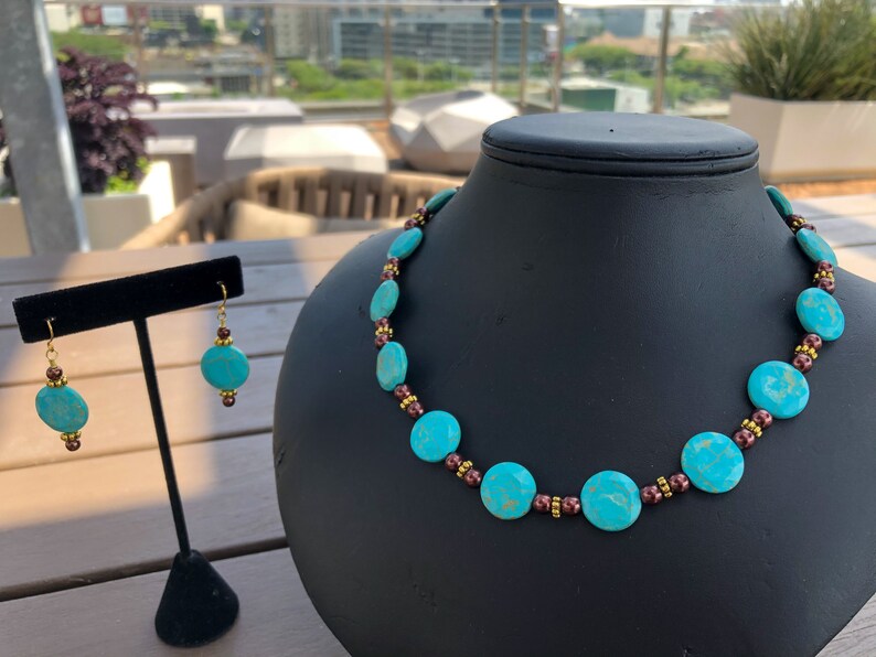 May include: A turquoise necklace and matching earrings set. The necklace features large, round turquoise beads interspersed with smaller brown and gold beads. The earrings have a similar design, with turquoise beads and gold hooks. The jewelry is displayed on a black mannequin.