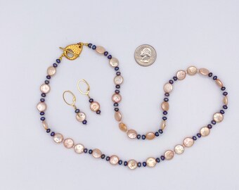 May include: A necklace and earring set featuring peach-colored, flat, round pearls and small, dark blue pearls. The necklace has a gold-colored clasp. The earrings have gold-colored hooks. A U.S. quarter is next to the jewelry.