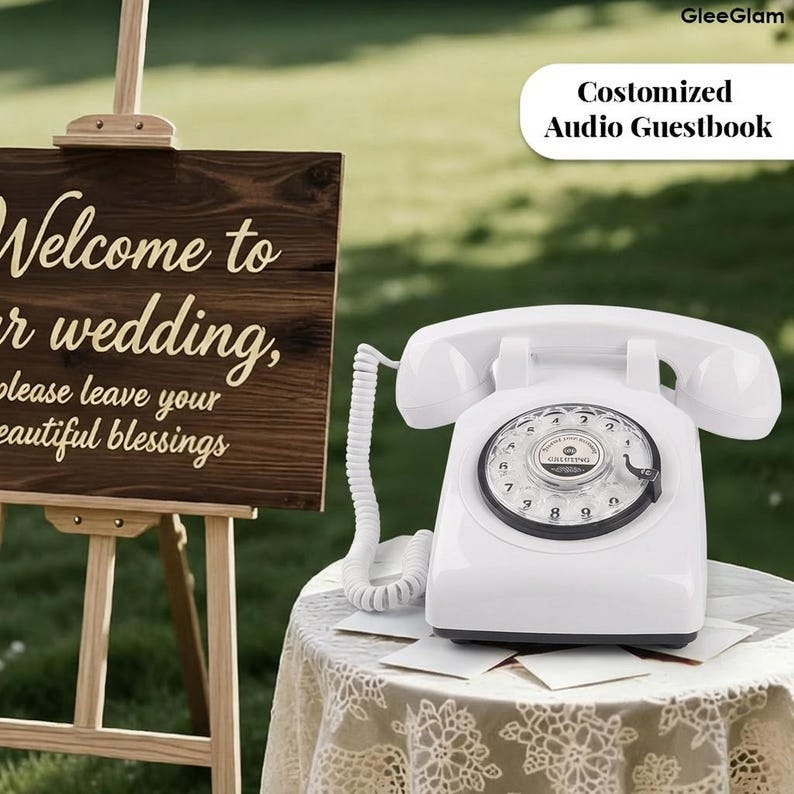May include: A white vintage rotary phone with a black dial sits on a lace-covered table. The phone is a wedding guestbook. A wooden sign with the words "Welcome to our wedding" is in the background.