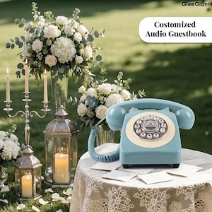 May include: A pale blue retro rotary phone with a cream dial on a lace-covered table. The scene includes white flowers, candles, and lanterns. The text reads "Customized Audio Guestbook". The setting suggests a wedding.