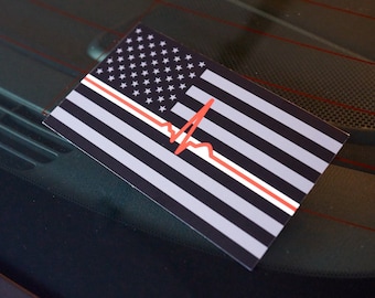 May include: Rectangular sticker featuring a stylized American flag design. The flag has a black and white striped pattern with a red line and a red heartbeat graphic. The sticker is placed on a car window.