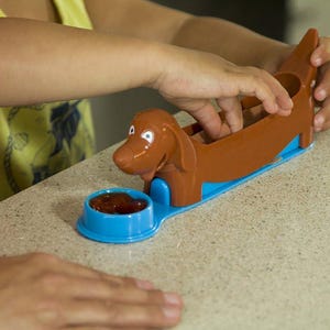 Puede incluir: Un cortador de perritos calientes de plástico marrón con forma de perro salchicha, con una base azul y un cuenco pequeño lleno de una sustancia roja. Una persona está manipulando el cortador.