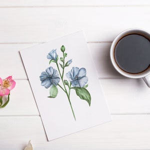 May include: A white card with a watercolor painting of blue flowers on a white wooden surface with a cup of coffee.