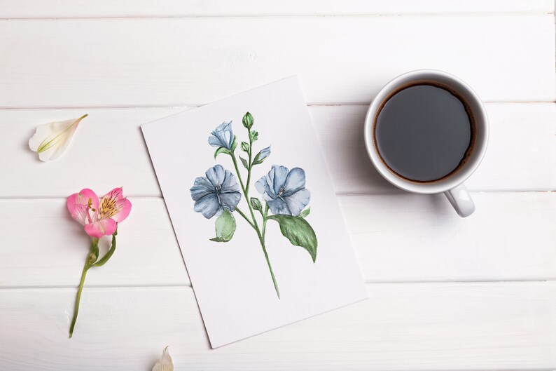 May include: A white card with a watercolor painting of blue flowers on a white wooden surface with a cup of coffee.