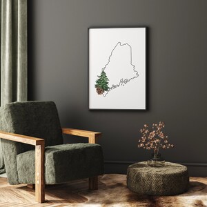 May include: A black framed print of the outline of the state of Maine with a small green pine tree and a brown pine cone in the lower left corner. The print is hanging on a gray wall above a green armchair and a brown woven ottoman.