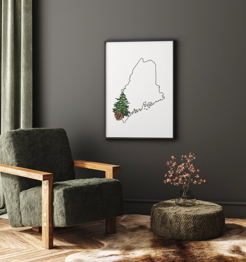 May include: A black framed print of the outline of the state of Maine with a small green pine tree and a brown pine cone in the lower left corner. The print is hanging on a gray wall above a green armchair and a brown woven ottoman.